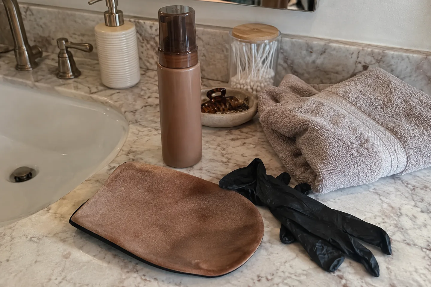 A self-tanning mousse bottle, a tanning mitt and a folded towel on a bathroom counter